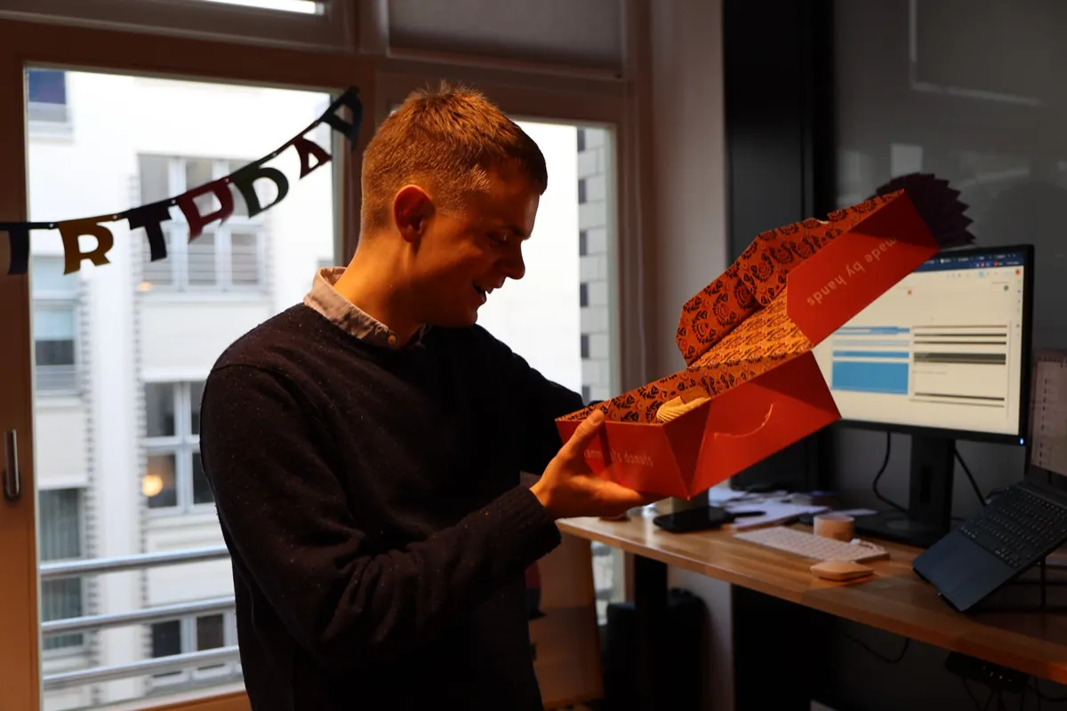 Johannes opening a red donut box at his desk, with a birthday garland in the window behind him