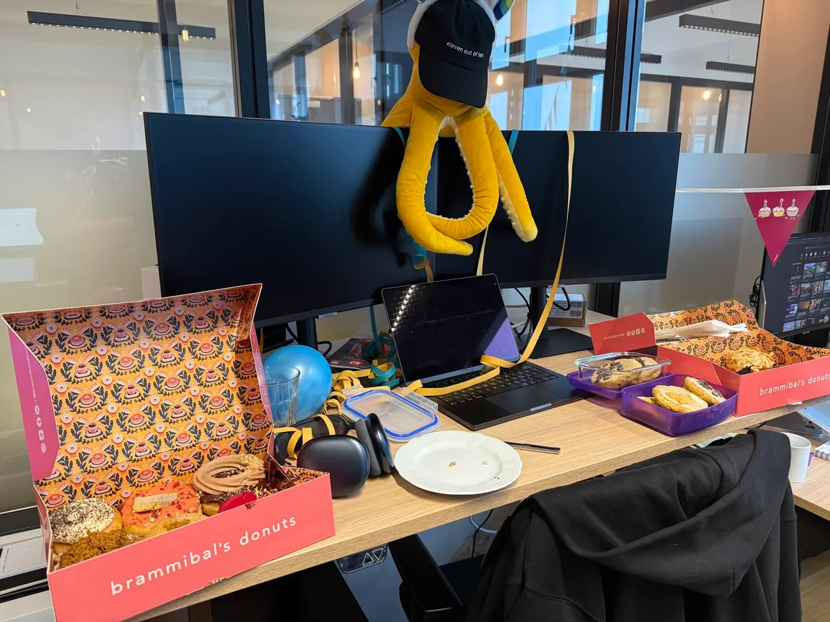 A desk with open boxes of donuts and a yellow octopus soft toy in a black cap hanging from the monitor