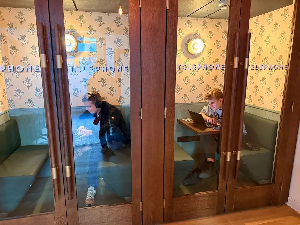 Johannes and Maxi working on laptops in wood-framed phone booths with patterned wallpaper