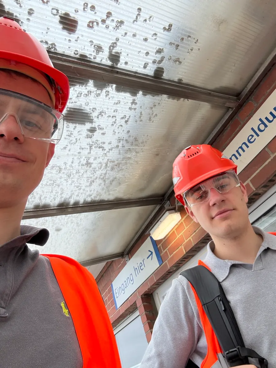 Selfie of Johannes and Maxi in red hard hats, safety glasses and high-visibility vests under a canopy at a plant entrance