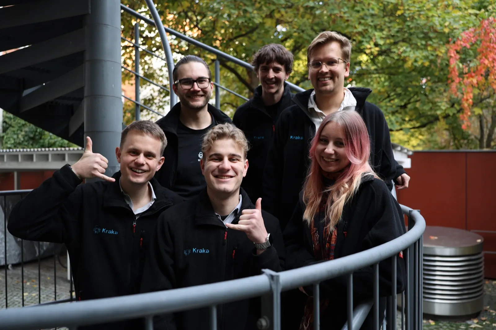 Six people from the Kraka team in black Kraka jackets on a spiral fire escape, smiling, two of them giving a thumbs up