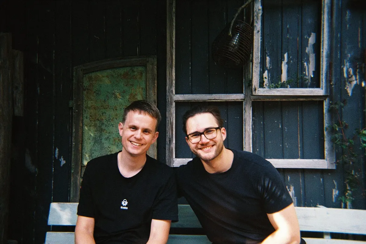 Johannes and Moritz in black T-shirts, smiling on a bench in front of a dark wooden wall